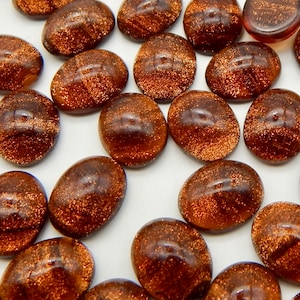May include: A collection of oval-shaped, brown glass cabochons with a sparkly, iridescent finish. The cabochons have a textured surface and are scattered on a white background.