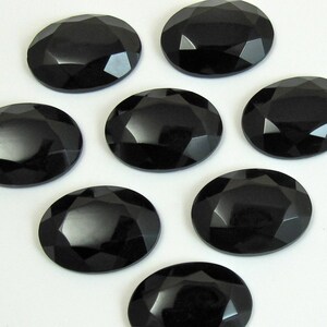May include: Seven black oval-shaped faceted gemstones. The gemstones have a high polish and are arranged on a white surface.