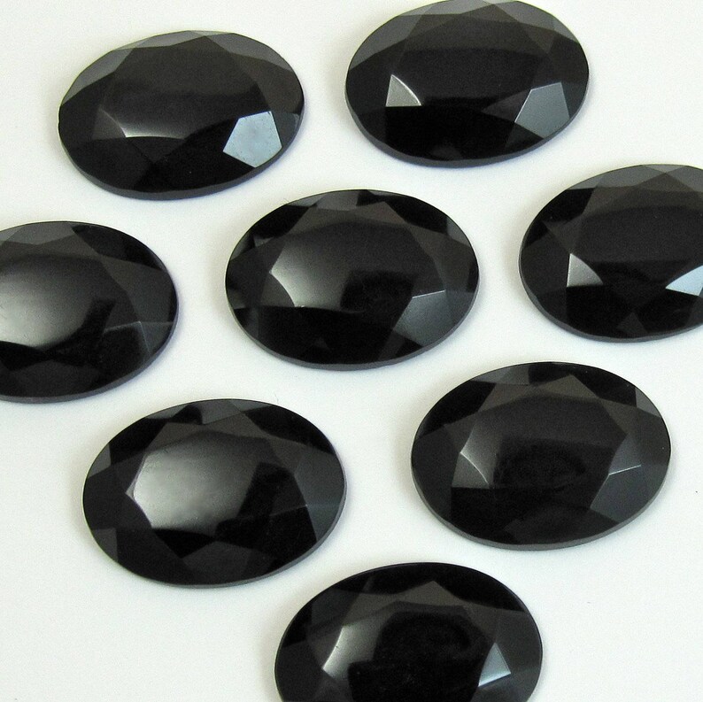 May include: Seven black oval-shaped faceted gemstones. The gemstones have a high polish and are arranged on a white surface.