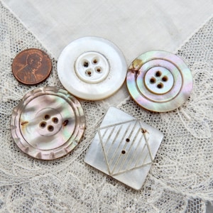 May include: A collection of vintage mother-of-pearl buttons in various shapes and sizes. Includes a round, white button, a circular button with iridescent hues, a scalloped button, and a diamond-shaped button. A U.S. penny is included for size reference.
