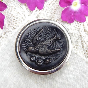 May include: A round, dark gray button with a detailed relief of a bird in flight, surrounded by wheat and floral elements. The button has a silver-colored metal rim. The background includes lace and purple flowers.