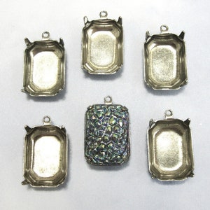 May include: Six silver-toned metal pendant settings with a beveled edge. The settings are rectangular in shape and have a loop at the top for attaching a chain or cord. One setting is filled with a iridescent blue and green glass cabochon.