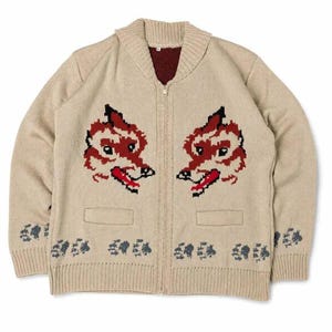 May include: A beige knit zip-up cardigan featuring a shawl collar and a dark red interior lining. The front displays two symmetrical red and black fox head motifs above two pockets, with a repeating dark gray paw print pattern along the hem.