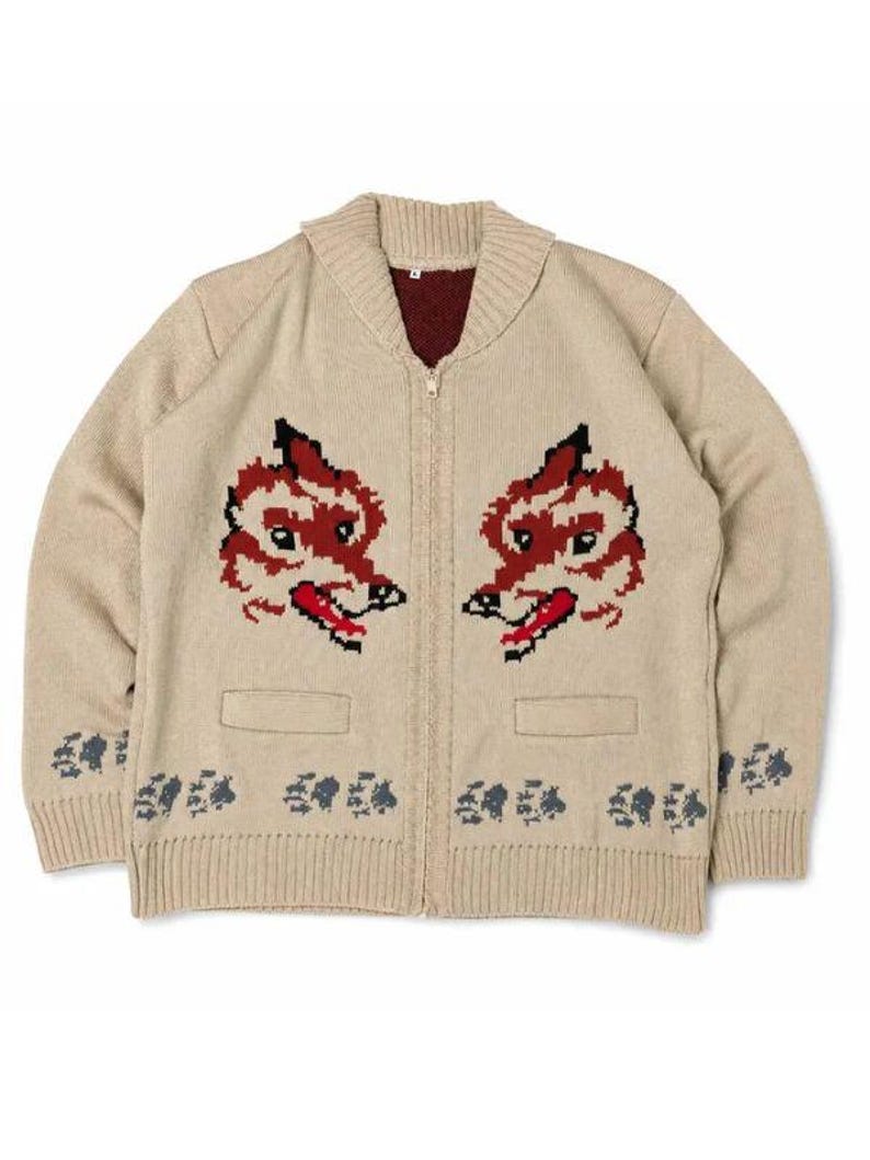 May include: A beige knit zip-up cardigan featuring a shawl collar and a dark red interior lining. The front displays two symmetrical red and black fox head motifs above two pockets, with a repeating dark gray paw print pattern along the hem.