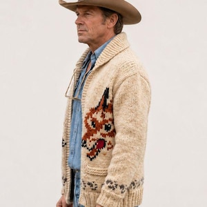 May include: A beige knit cardigan featuring a shawl collar and a large fox graphic on the front. The sweater is worn over a light blue button-down shirt and blue jeans, paired with a tan felt cowboy hat and glasses hanging from the shirt collar.