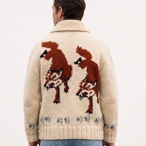 May include: A cream-colored knit cardigan featuring a shawl collar and a back design of two rust-orange foxes. The hem and cuffs are decorated with a repeating pattern of small grey paw prints. The garment is worn over light blue denim jeans.