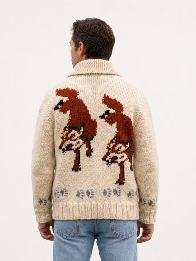 May include: A cream-colored knit cardigan featuring a shawl collar and a back design of two rust-orange foxes. The hem and cuffs are decorated with a repeating pattern of small grey paw prints. The garment is worn over light blue denim jeans.