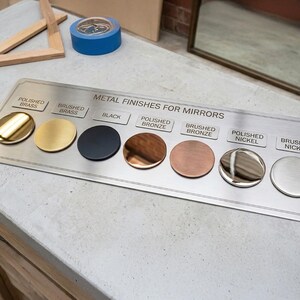 Może przedstawiać: Prostokątna metalowa płytka wzornikowa z napisem METAL FINISHES FOR MIRRORS, zawierająca osiem okrągłych pr&oacute;bek o r&oacute;żnych wykończeniach: polerowany mosiądz, szczotkowany mosiądz, czarny, polerowany brąz, szczotkowany brąz, polerowany nikiel i szczotkowany nikiel.