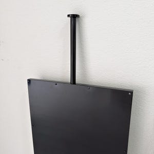 Może przedstawiać: Czarny, trapezoidalny panel ekspozycyjny o gładkiej, odblaskowej powierzchni. Pionowy, cylindryczny czarny pręt wystaje w g&oacute;rę ze środka g&oacute;rnej krawędzi i jest zakończony małą, okrągłą, płaską nasadką. Panel jest przymocowany za pomocą śrub.