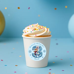 Puede incluir: Una taza de papel blanca con una magdalena cubierta con glaseado blanco y chispas de colores. La taza tiene una pegatina con el número 3, copos de nieve y personajes de la película Frozen. Un globo dorado y un globo azul en el fondo.
