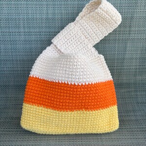 May include: A crochet handbag featuring a color-blocked design with horizontal stripes of white, orange, and yellow. The bag has a single integrated handle formed by one side being longer than the other, creating a loop closure style.