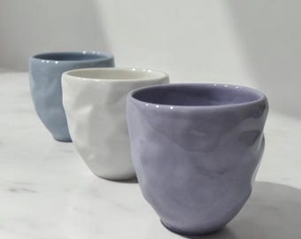 May include: Three ceramic cups in different shades of blue and white. The cups have a textured, crumpled appearance.
