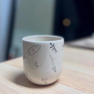 Puede incluir: Una taza de cerámica blanca con dibujos lineales negros de una hogaza de pan, una ramita, una vela, una luna creciente, un corazón, un adorno y una copa de cóctel. La taza tiene forma redondeada y un acabado liso y brillante.