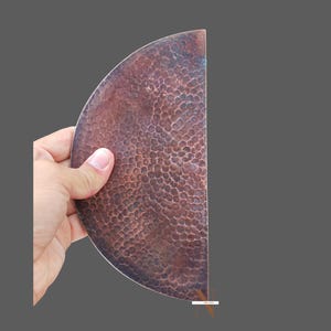 May include: A half-circle copper plate with a hammered texture, held in a hand. The plate's surface is a rich, reddish-brown with a dimpled pattern. The edges are smooth, and the background is a solid grey. The plate is likely for decorative use.