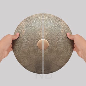 May include: Two-piece, round, bronze-colored door handles with a textured surface and a central, circular, brass-colored accent. The handles are held open against a neutral background, showcasing their design and finish. Ideal for home decor.