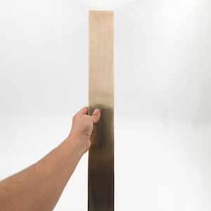 May include: A rectangular, brass-coloured metal bar with a gradient finish, held against a white background. The bar transitions from a lighter gold at the top to a darker, bronze hue at the bottom. The object is held by a hand.