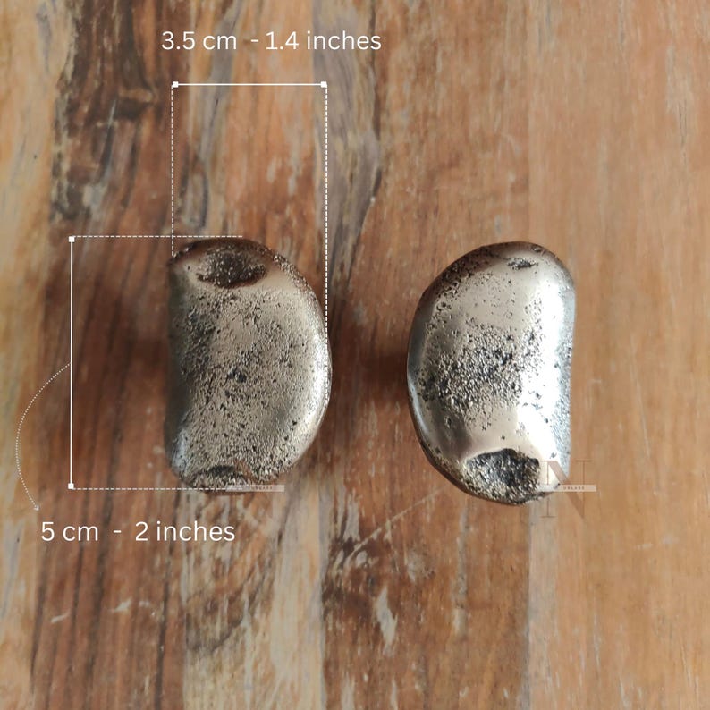 Puede incluir: Dos pomos met&aacute;licos para armarios, cada uno de aproximadamente 5 cm de largo y 3,5 cm de ancho. Los pomos tienen una superficie texturizada y moteada y una forma redondeada y org&aacute;nica. Se muestran sobre una superficie de madera.