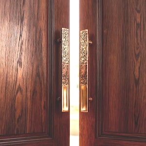 Puede incluir: Un par de puertas de madera oscura con manijas doradas ornamentadas. Las manijas son rectangulares con patrones de relieve detallados. Las puertas están ligeramente entreabiertas, revelando una luz brillante entre ellas. La veta de la madera es visible.