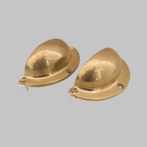 May include: Two brass-colored, shell-shaped drawer pulls. Each pull has a rounded, curved body and a flat base with screw holes. The pulls have a smooth, polished finish and are designed for furniture hardware.