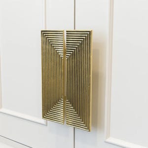 pull with a geometric design. The door pull features a series of recessed, vertical lines and a pattern of concentric squares. The surface has a textured appearance, and the overall design is modern and decorative. The door pull is held against a white background.