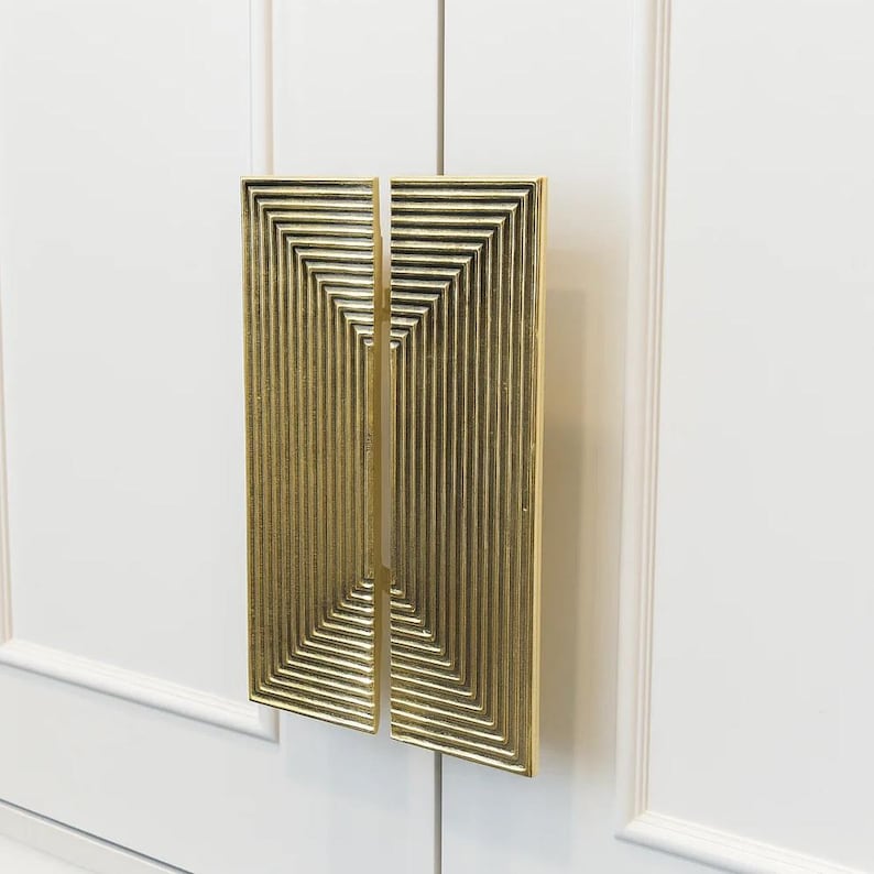 pull with a geometric design. The door pull features a series of recessed, vertical lines and a pattern of concentric squares. The surface has a textured appearance, and the overall design is modern and decorative. The door pull is held against a white background.
