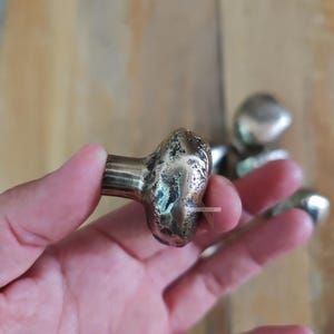 Puede incluir: Primer plano de un pomo de armario de color bronce con un diseño orgánico texturizado. El pomo tiene una base cilíndrica y una parte superior redondeada e irregular. Se ven pomos adicionales en el fondo, lo que sugiere un juego para herrajes de muebles.