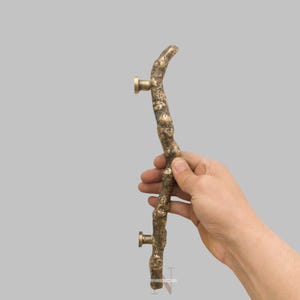 Pode incluir: Uma maçaneta de porta bronzeada com um design de galho orgânico texturizado. A maçaneta tem um formato curvo e é montada com duas fixações redondas de cor latão. A estética geral é rústica e natural.