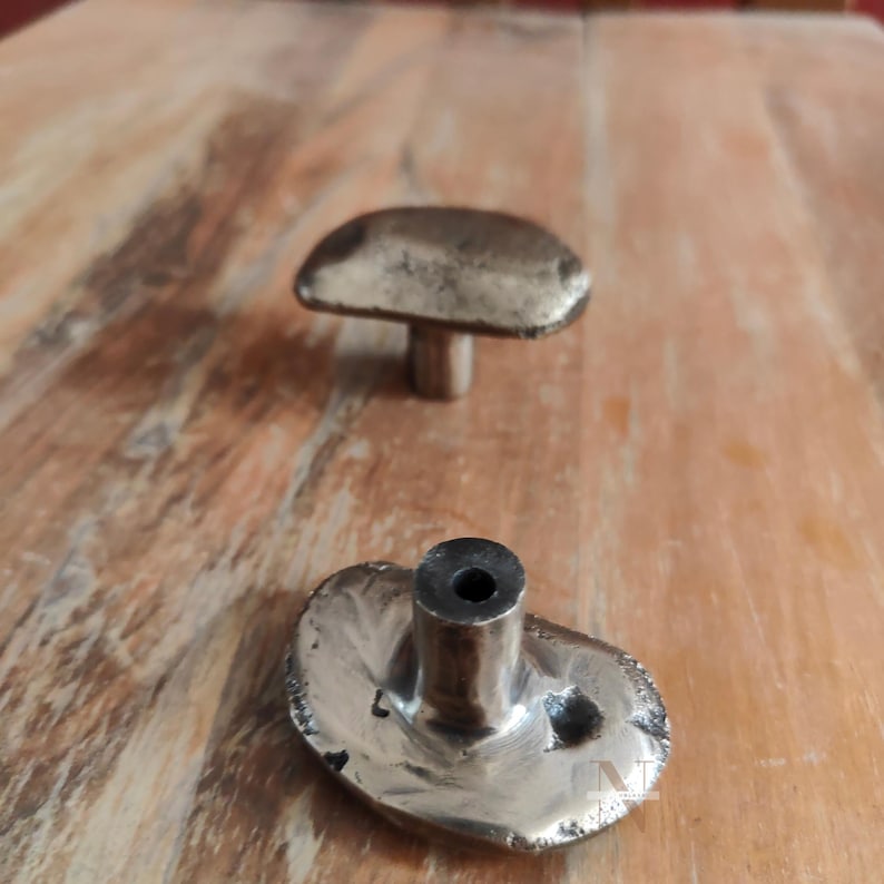 Puede incluir: Dos tiradores de armario antiguos con forma de hongo de bronce. Los tiradores tienen una superficie texturizada y un v&aacute;stago para el montaje. Los sombreros de los hongos son ovalados y ligeramente abovedados. Los tiradores est&aacute;n sobre una superficie de madera marr&oacute;n claro.