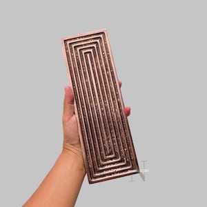 Könnte beinhalten: Ein rechteckiger, kupferfarbener Tablett mit einem vertieften, labyrinthartigen Design. Das Tablett wird in einer Hand gehalten, wodurch das komplizierte Muster und die metallische Oberfläche hervorgehoben werden. Das Design weist parallele Linien und rechte Winkel auf.