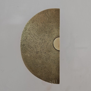 May include: A half-circle brass door pull with a textured surface and a small, round accent. The hardware is a warm gold color and is mounted on a white surface. The design is modern and minimalist.