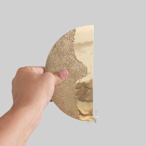 Puede incluir: Un objeto decorativo dorado en forma de semicírculo, con un patrón texturizado y punteado en un lado y una superficie lisa y reflectante en el otro. El objeto se sostiene sobre un fondo gris.