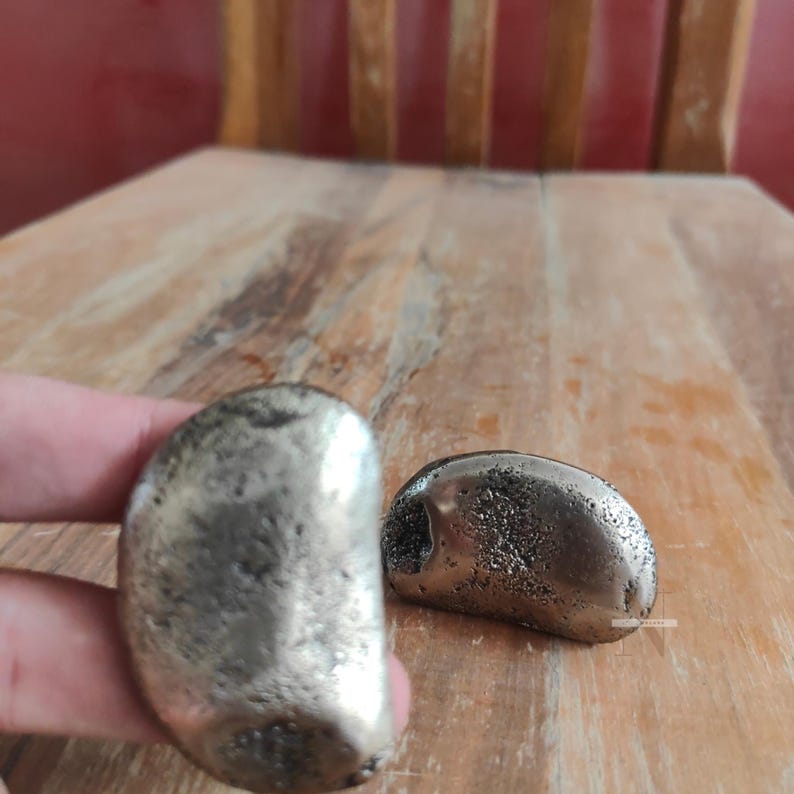 Puede incluir: Dos objetos decorativos plateados en forma de guijarro. Uno est&aacute; sostenido por una mano, el otro descansa sobre una superficie de madera. Los objetos tienen una superficie texturizada y picada, y una forma suave y redondeada. El fondo es una mesa de madera y una pared roja.