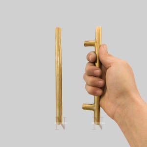 Könnte beinhalten: Zwei messingfarbene Schrankgriffe. Einer ist ein gerader Stab, der andere wird in einer Hand gehalten und hat eine T-Form. Die Griffe haben eine strukturierte Oberfläche und werden vor einem hellgrauen Hintergrund gezeigt. Der gerade Stabgriff ist ca. 25 cm lang.