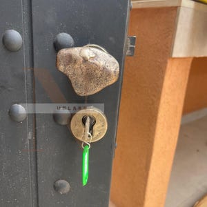 Op de afbeelding: Close-up van een zwarte metalen deur met een decoratieve, getextureerde bronzen handgreep en sleutelgat. Een sleutel met een groen label zit in het slot. De deur heeft grote, ronde, donkere metalen klinknagels.