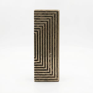 May include: A rectangular, bronze-colored object with a geometric design. The design consists of a series of concentric rectangles that create a textured surface. The object is likely a decorative piece or a small sculpture.