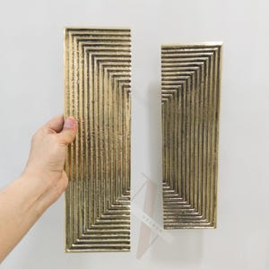Può includere: Due maniglie per porte rettangolari color oro con un design geometrico scanalato. Le maniglie sono tenute su uno sfondo bianco. Il design presenta una serie di rettangoli concentrici, creando una superficie strutturata.