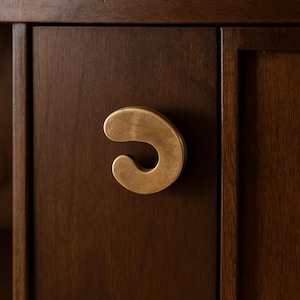 Puede incluir: Primer plano de una puerta de armario con un tirador curvo de color latón. El tirador tiene una forma orgánica única y está montado en un armario de madera marrón oscuro. La veta de la madera es visible, añadiendo textura.