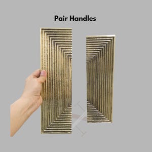Może przedstawiać: Para złotych, prostokątnych uchwyt&oacute;w o geometrycznym wzorze. Uchwyty mają szereg koncentrycznych kwadrat&oacute;w i pionowych linii. Tekst "Pair Handles" znajduje się u g&oacute;ry obrazu.