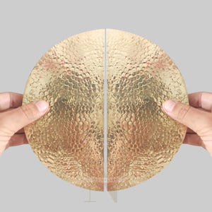 May include: Two halves of a gold-colored, hammered-metal, semi-circular door pull. The textured surface has a dimpled pattern. The door pulls are held by hands against a light gray background. The pulls are designed to be installed together to form a complete circle.