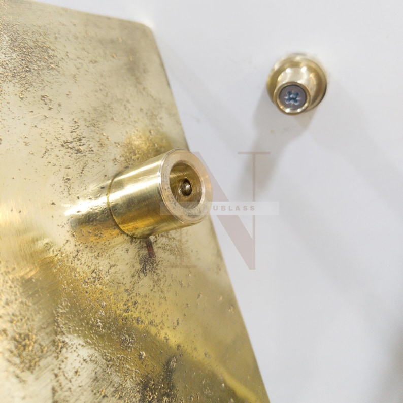 Może przedstawiać: Zbliżenie mosiężnego elementu z cylindrycznym elementem i łbem śruby. Mosiądz ma teksturowany, postarzany wygląd. Element jest zamontowany na białej powierzchni.