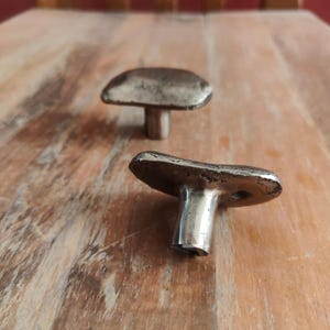 Puede incluir: Dos tiradores de armario de metal de estilo antiguo con un diseño en forma de hongo. Los tiradores son de color plateado con un aspecto ligeramente deslustrado y están colocados sobre una superficie de madera cálida. Los tiradores son probablemente para muebles o decoración.