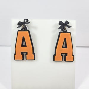 May include: A pair of orange letter "A" earrings with black trim and small black bows at the top. The earrings are displayed on a white surface, showcasing their bold design and color contrast. These earrings are a fun accessory.