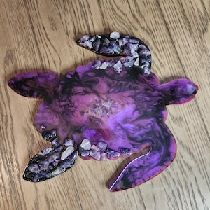 May include: A purple and black resin turtle-shaped tray with a swirling design. The tray is adorned with small, irregularly shaped, light-colored stones along the edges of the flippers, head, and tail.