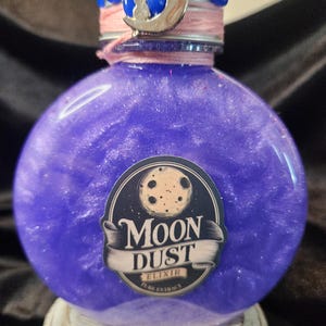 May include: A round glass bottle filled with a shimmering purple liquid. A label on the front features a moon illustration and the text MOON DUST ELIXIR PURE EXTRACT. The bottle cap is sealed with blue wax, pink thread, and a silver crescent moon charm.