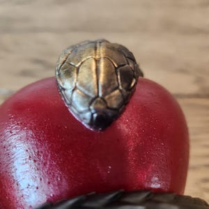 May include: A detailed close-up of a snake-themed decorative object. The focal point is the metallic, textured head of a snake, positioned atop a smooth, red, rounded form. The snake's head is a mix of gold and bronze tones, with a dark, textured body.