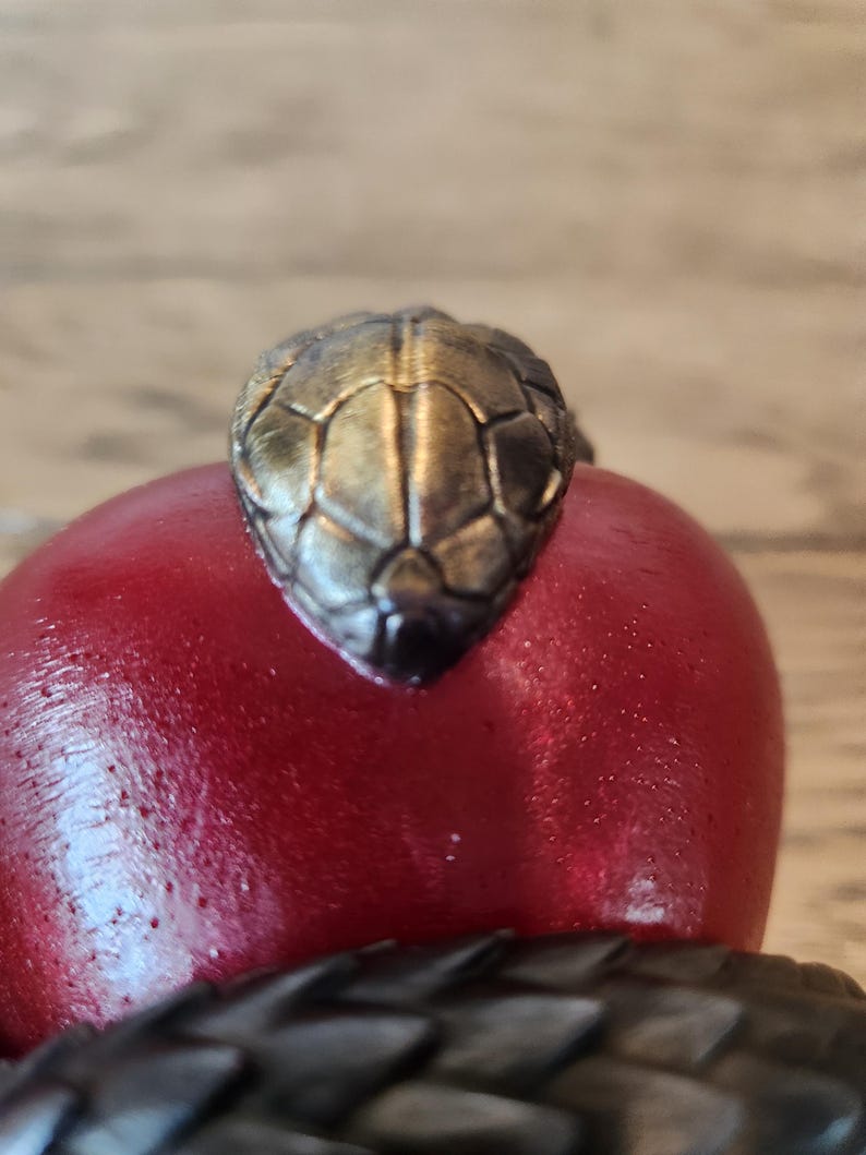 May include: A detailed close-up of a snake-themed decorative object. The focal point is the metallic, textured head of a snake, positioned atop a smooth, red, rounded form. The snake's head is a mix of gold and bronze tones, with a dark, textured body.
