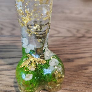 Może przedstawiać: Przezroczysty kieliszek do shot&oacute;w z żywicy ze złotymi płatkami, mosiężnym kluczem i złotą pszczołą. Podstawa jest zielona z białymi kwiatami i mchem. Kieliszek do shot&oacute;w stoi na drewnianej powierzchni.