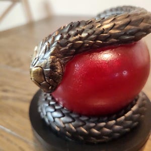 May include: A sculpture featuring a bronze-colored snake coiled around a red sphere. The snake's head is at the top, with detailed scales and a textured appearance. The sculpture sits on a black base.