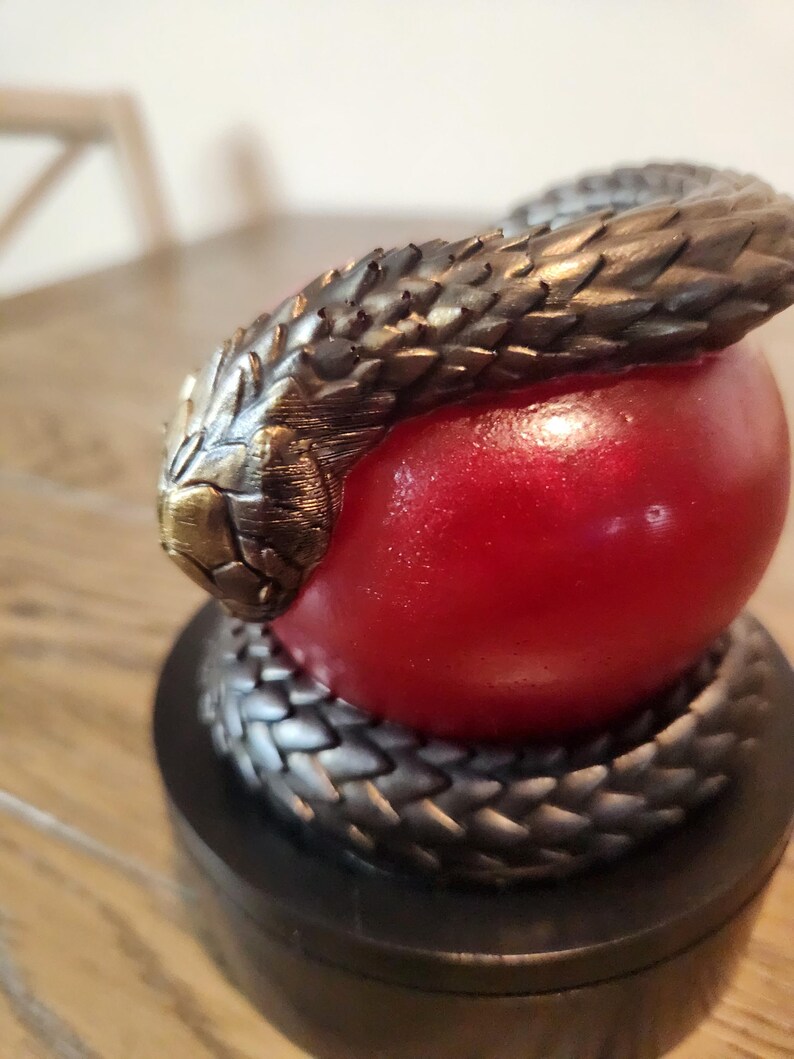 May include: A sculpture featuring a bronze-colored snake coiled around a red sphere. The snake's head is at the top, with detailed scales and a textured appearance. The sculpture sits on a black base.
