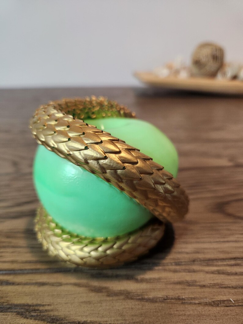 May include: A gold-colored, textured snake wraps around a light green sphere. The snake's scales are detailed, and the sphere has a smooth surface. The design is set against a blurred background.